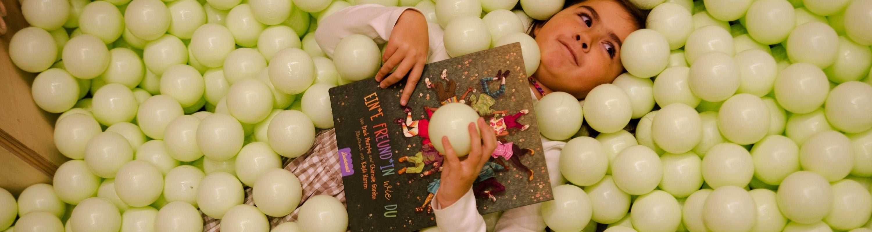 Ein Kind liegt in einer mit blassgrünen Bällen gefüllten Grube, hält ein Buch in der Hand und schaut lächelnd nach oben.