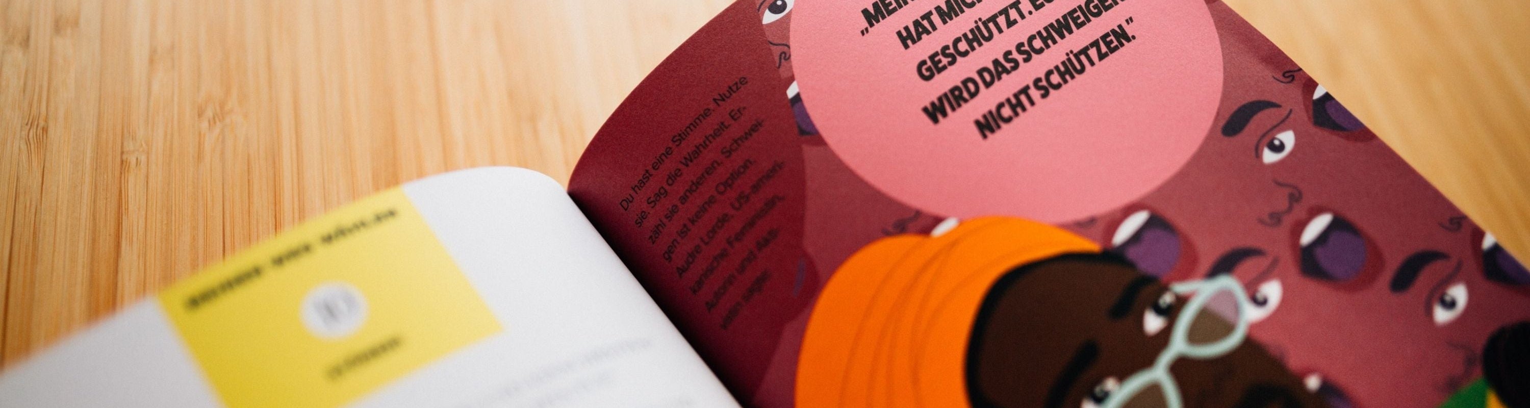 Ein aufgeschlagenes Buch auf einer hölzernen Oberfläche, das bunte Illustrationen von Gesichtern und deutschen Text in einer Sprechblase auf der rechten Seite zeigt. Die linke Seite enthält einen gelben Abschnitt mit Text.