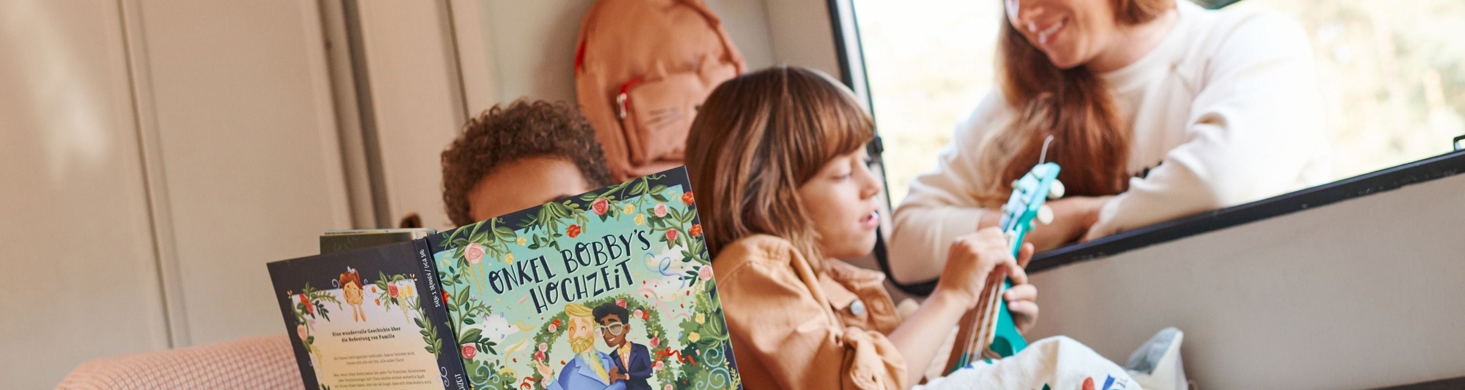 Zwei Kinder sitzen auf einem Sofa; eines liest ein buntes Buch mit dem Titel "Onkel Bobbys Hochzeit", während das andere eine kleine blaue Gitarre spielt. Eine Erwachsene lächelt ihnen durch ein nahes Fenster zu.