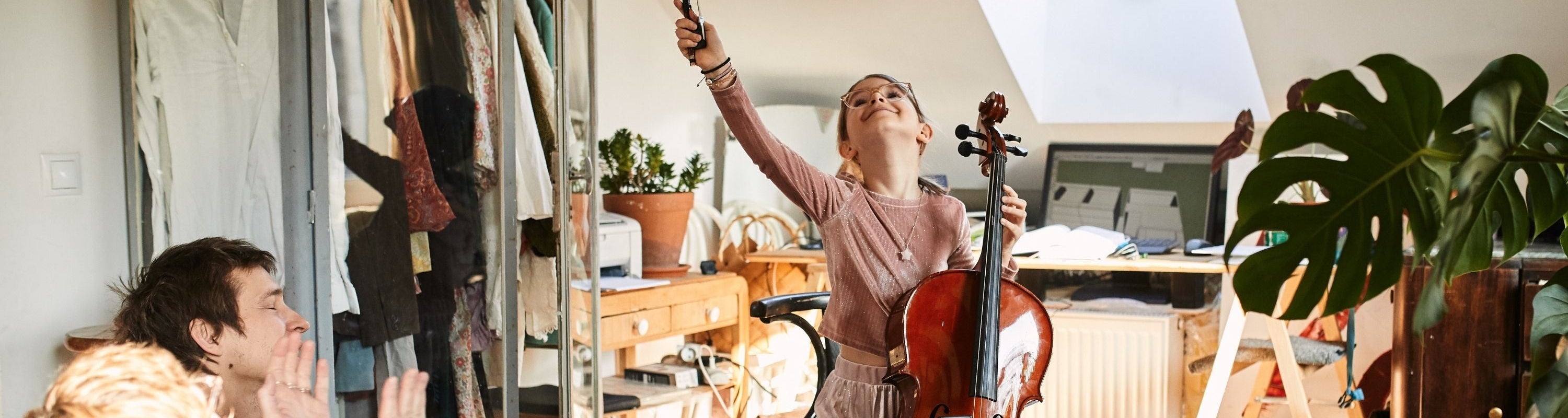 Ein junges Mädchen spielt in einem hellen Raum begeistert Cello und hebt den Bogen in die Luft, während Erwachsene in der Nähe zusehen und klatschen. Der Raum ist gemütlich mit Pflanzen, Kleidern und einfallendem Sonnenlicht.