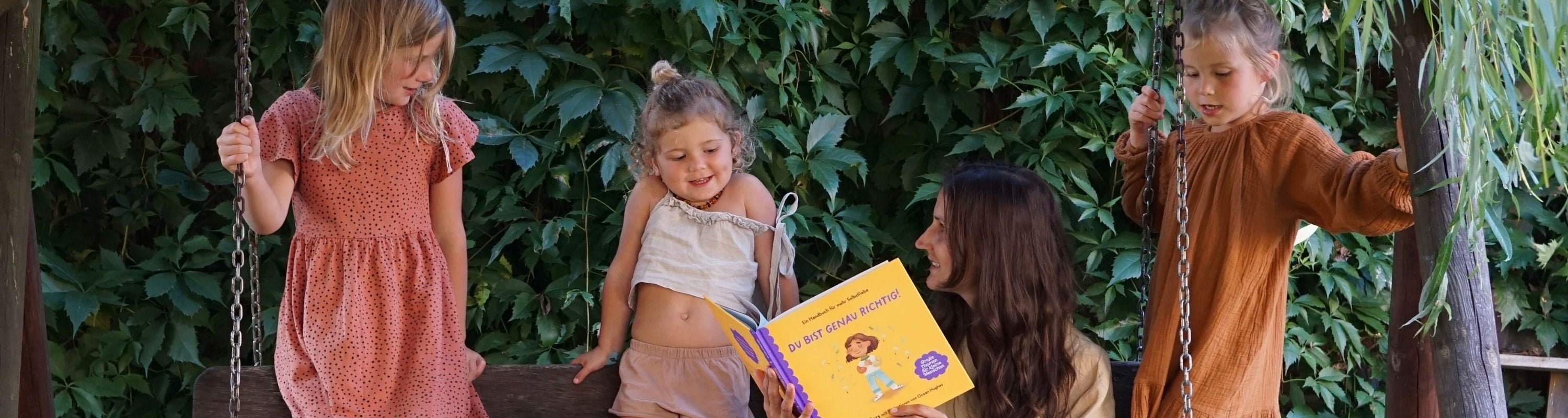 Eine Frau liest drei jungen Mädchen auf einer hölzernen Schaukelbank im Freien ein Kinderbuch vor, mit grünen Blattpflanzen im Hintergrund. Zwei Mädchen stehen auf der Schaukel, und eines sitzt auf der Bank.