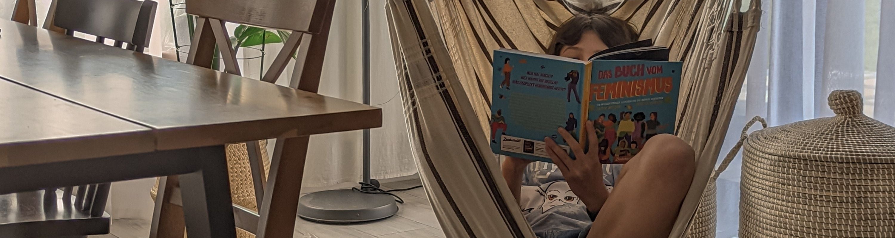 Ein Kind entspannt sich in einem Hängesessel im Haus und liest ein illustriertes Buch. Das Sonnenlicht strömt durch die durchsichtigen Vorhänge, in der Nähe stehen ein Holztisch und Stühle.