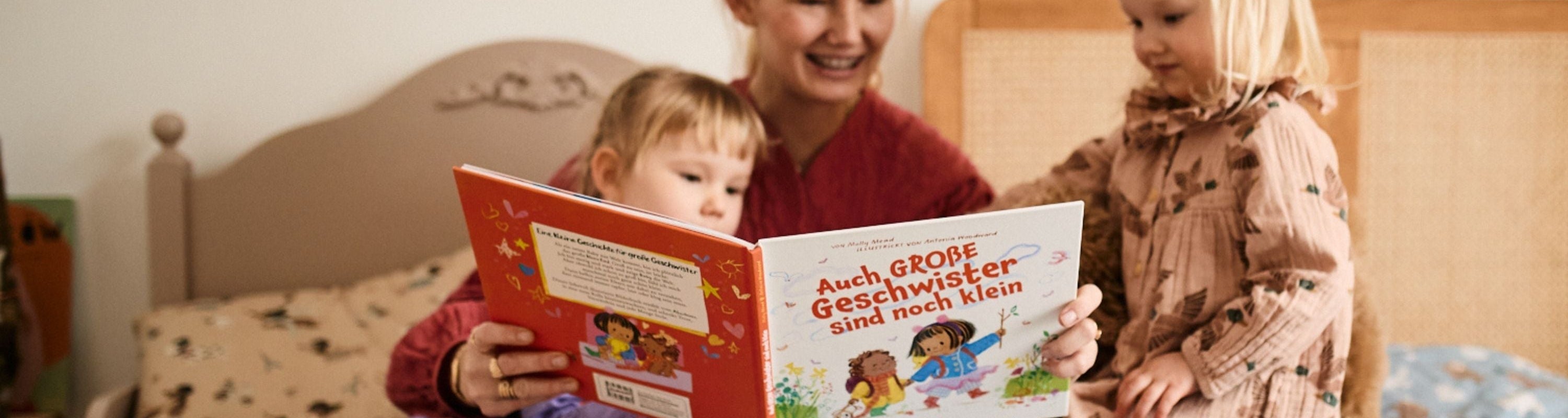 Eine Frau sitzt auf einem Bett und liest zwei jungen Mädchen aus einem deutschen Kinderbuch vor. Ein Mädchen sitzt neben ihr, das andere steht, beide schauen in das Buch und lächeln.