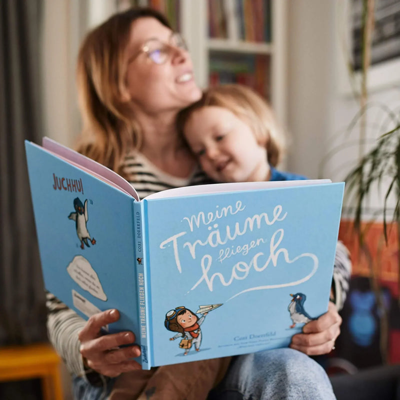 Eine Frau und ein kleines Kind lächeln, während sie ein deutsches Kinderbuch mit dem Titel Meine Träume fliegen hoch lesen. Der blaue Einband zeigt einen Pinguin und ein Kind mit Papierflieger