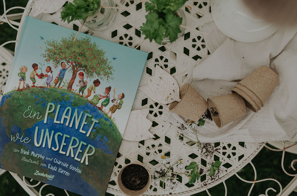 Ein deutsches Kinderbuch mit dem Titel Ein Planet wie unserer liegt auf einem weißen, verzierten Tisch neben verstreuter Erde, kleinen Pflanzentöpfen und grünen Blättern, die auf eine Gartenarbeit hindeuten.