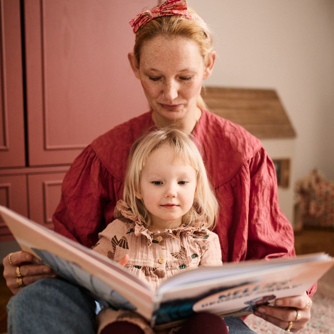 Eine Frau in roter Bluse und Stirnband liest einem kleinen Kind auf dem Schoß "Nellies großer Sprung" vom Zuckersüß Verlag - ein Kinderbuch über Mut und innere Stärke - vor, während im Hintergrund rote Schränke und ein kleines hausförmiges Objekt zu sehen sind.
