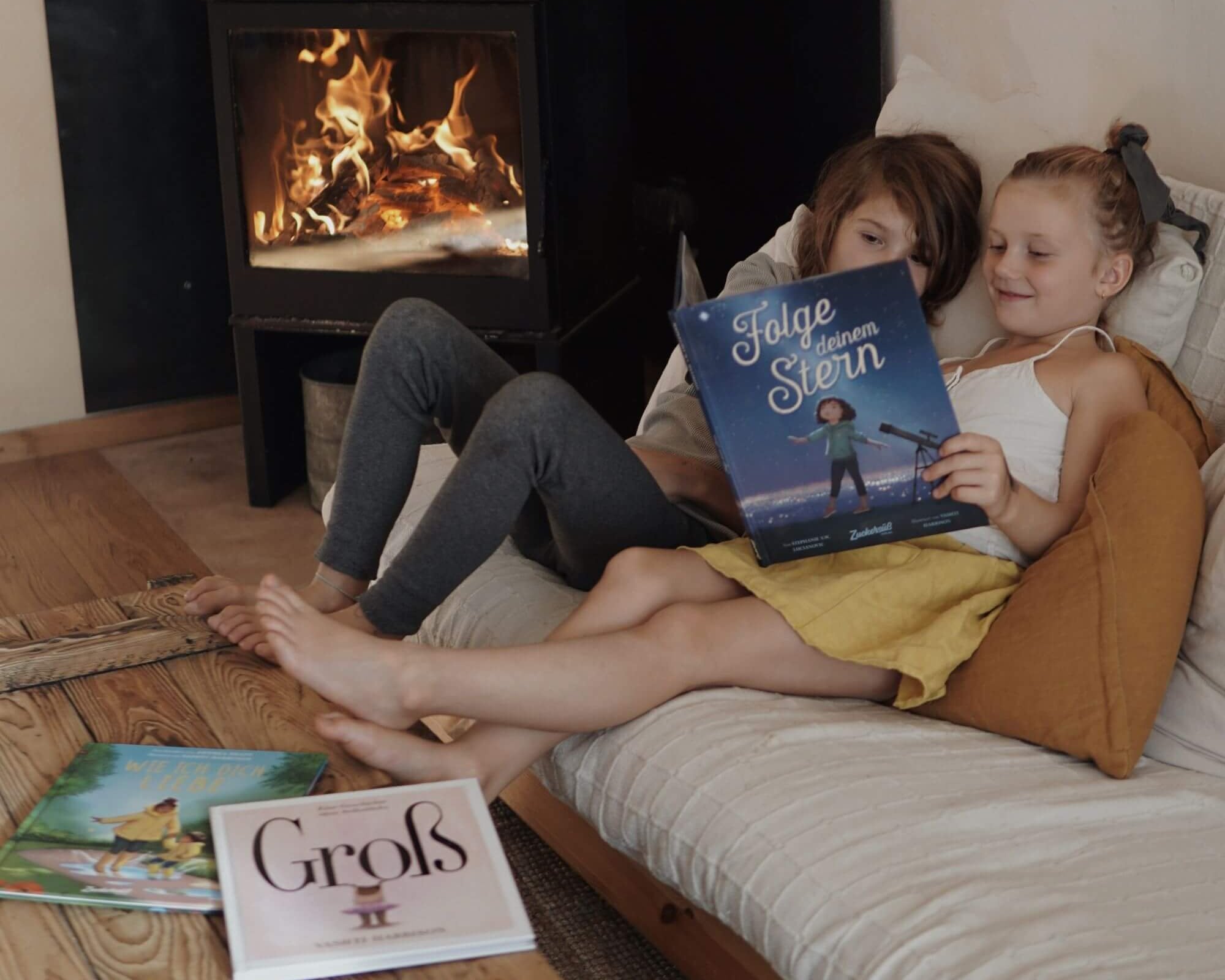Zwei Kinder sitzen auf einer Couch am Kamin in einem gemütlichen Kinderzimmer und lesen "Best of Vashti Harrison" aus dem Zuckersüß Verlag. Zwei weitere herzerwärmende Bücher liegen auf dem Boden und machen die Szene gemütlich und warm.