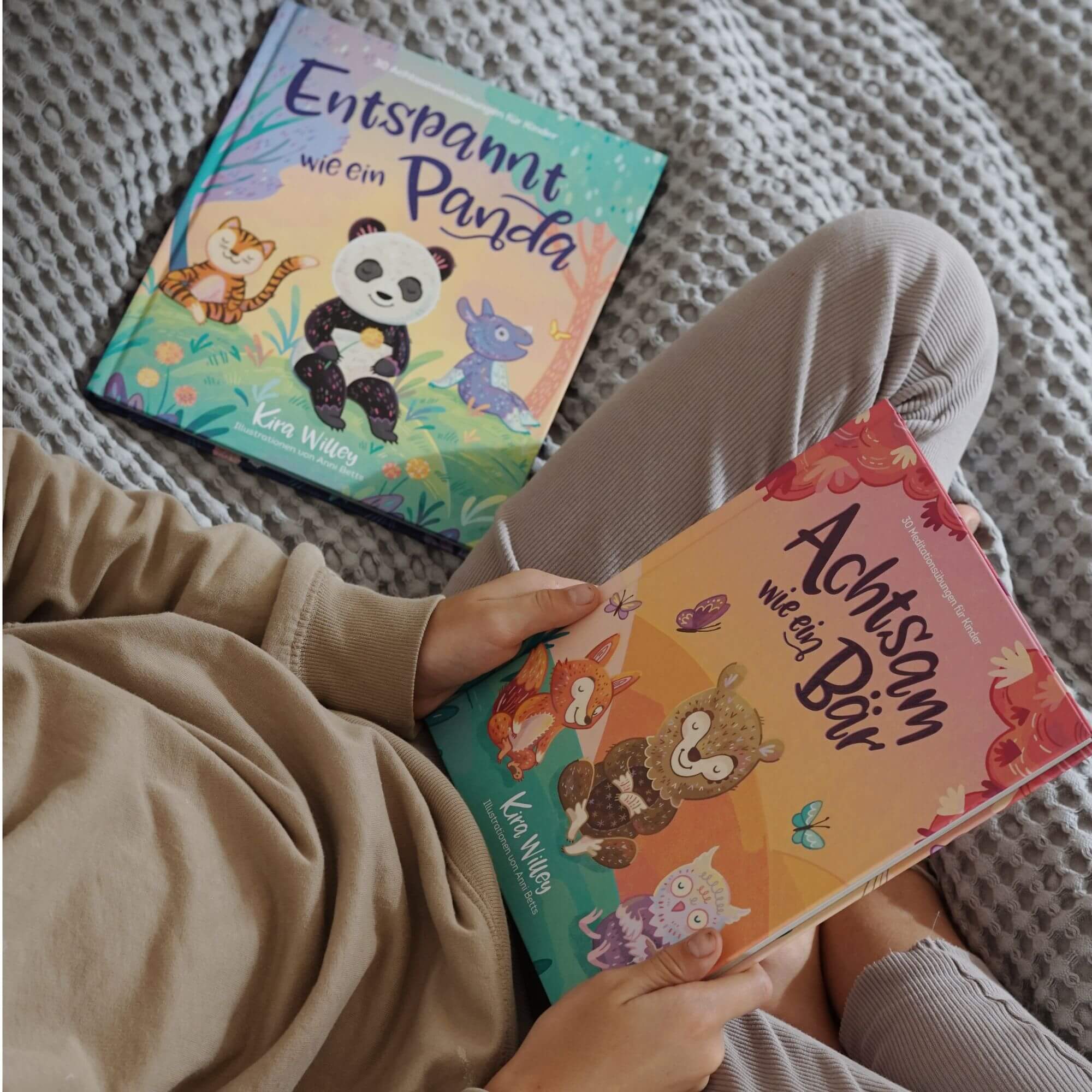 Ein Kind in beigefarbener Kleidung sitzt auf einer grauen Decke und hält das Buch „Achtsam wie ein Bär“ aus dem Doppelset „Achtsamkeit im Doppelpack“ des Zuckersüß Verlags in den Händen. Daneben liegt das zweite Buch der Reihe, „Entspannt wie ein Panda“. Beide Titel zeigen liebevoll illustrierte Tiere.