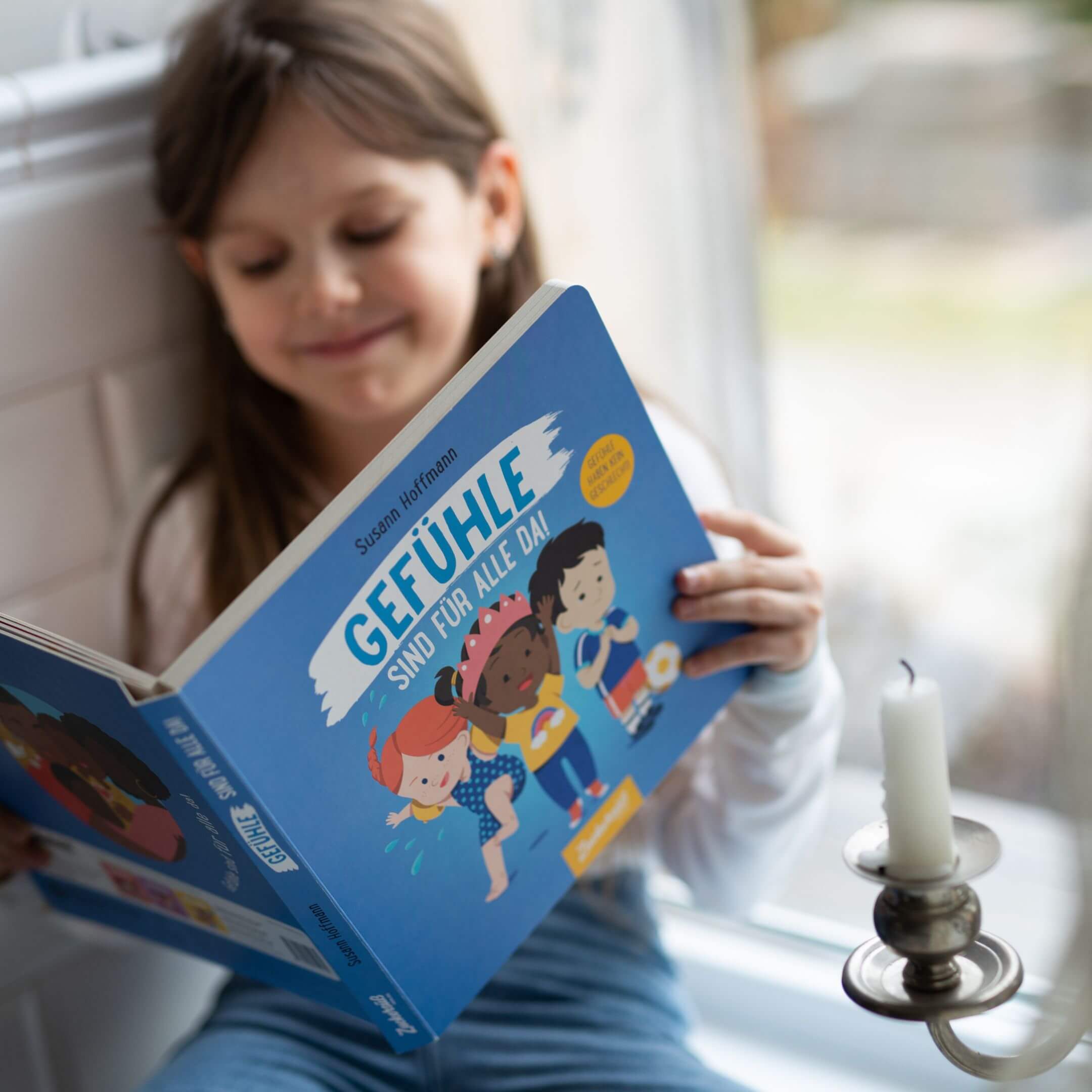 Ein junges Kind lächelt an einem Fenster, während es "Gefühle sind für alle da!" von Susann Hoffmann liest, ein Kinderbuch über Gefühle und integrative Werte. Im Vordergrund wird eine weiße Kerze auf einem Halter angezündet.