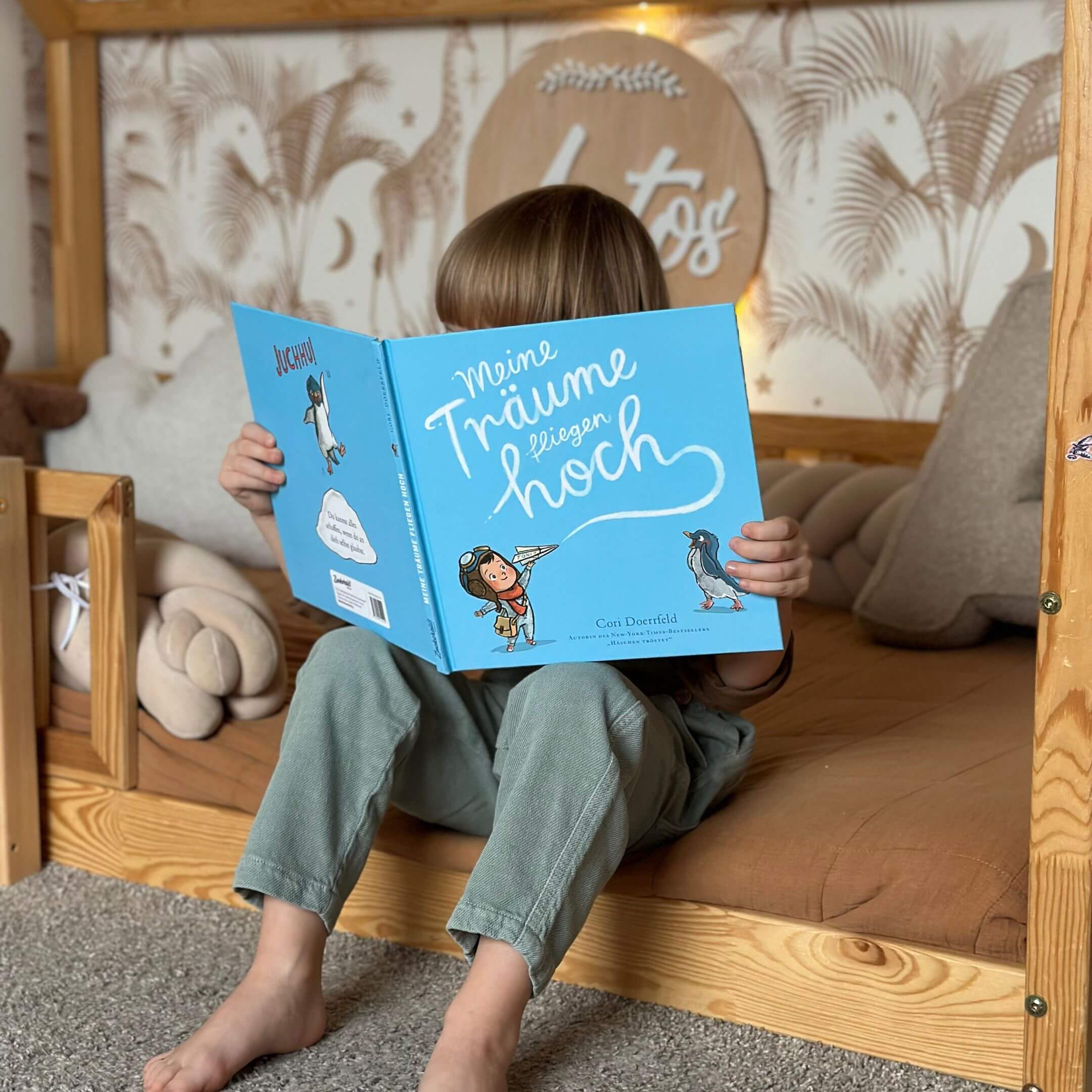 Ein Kind sitzt auf einem Holzbett und hält das Buch "Meine Träume fliegen hoch" von Cori Doerrfeld vor das Gesicht. Umgeben von Kuscheltieren, Deko Palmen und einem Namensschild.