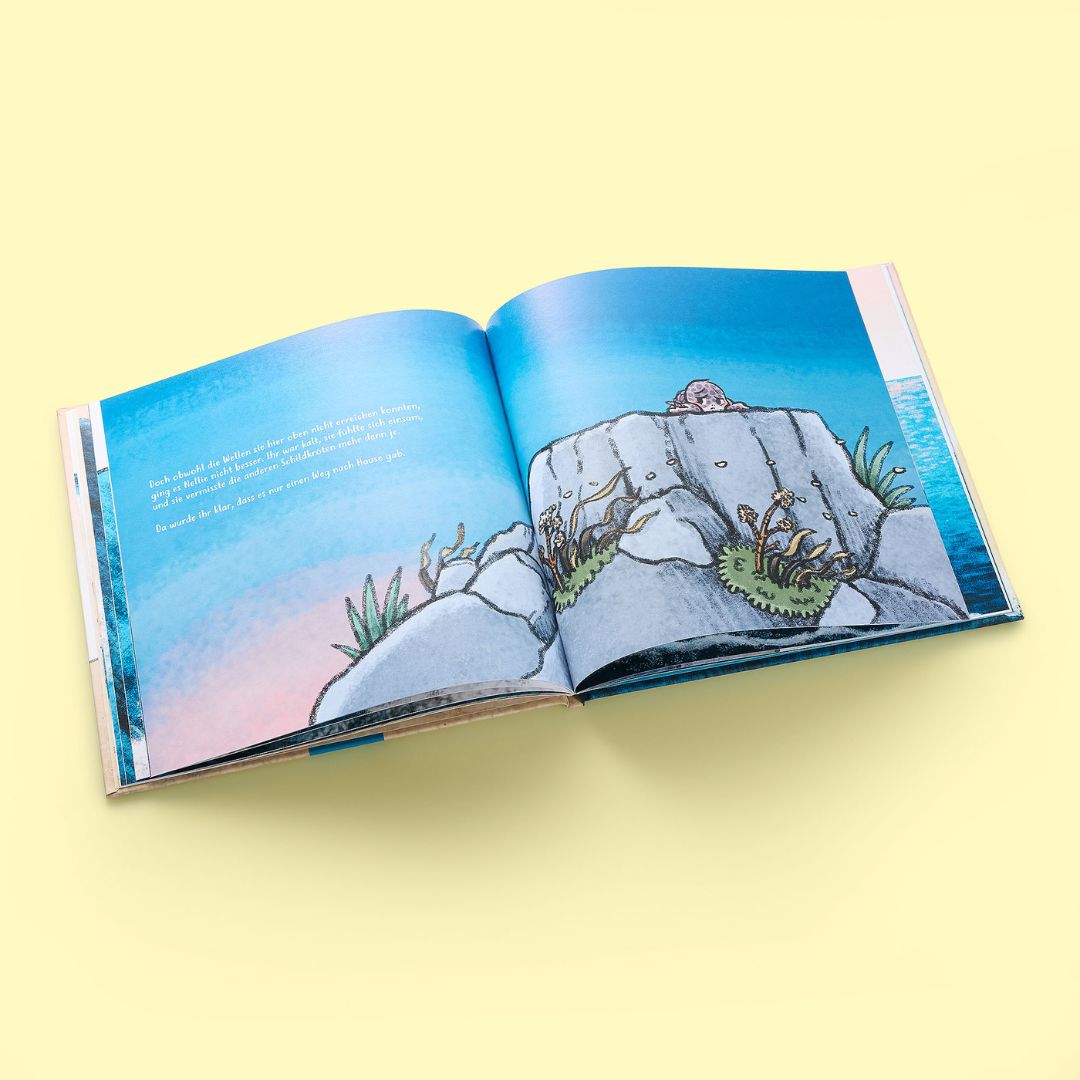 Ein aufgeschlagenes Exemplar von "Nellies großer Sprung" aus dem Zuckersüß Verlag liegt auf einer gelben Fläche und zeigt eine lebendige Szene mit einem Felsvorsprung, Seetang und einem Tier unter einem blauen Himmel mit Farbverlauf.