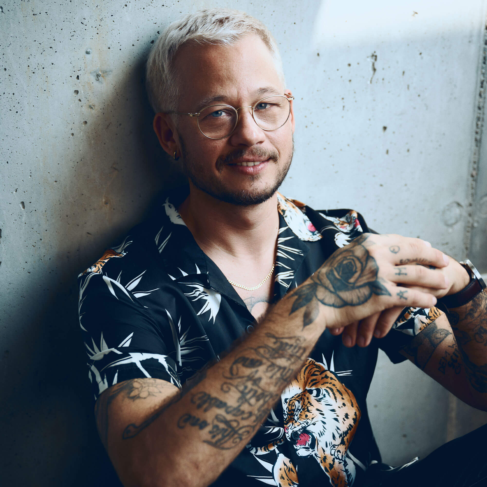 Henri Maximilian Jakobs sitzt mit seinem Buch "All die brennenden Fragen" an einer Betonwand und lächelt sanft in ihrem auffällig gemusterten Hemd und den platinblonden Haaren.