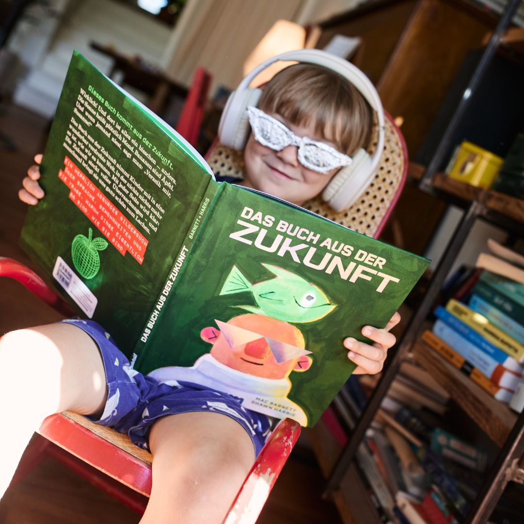 Ein Kind mit Kopfhörern und einer silbernen Spielzeugbrille sitzt auf einem Stuhl, lächelt und liest das deutsche Kinderbuch „Das Buch aus der Zukunft“ vom Zuckersüß Verlag in einem gemütlichen Zimmer, hinter dem sich Bücherregale befinden.