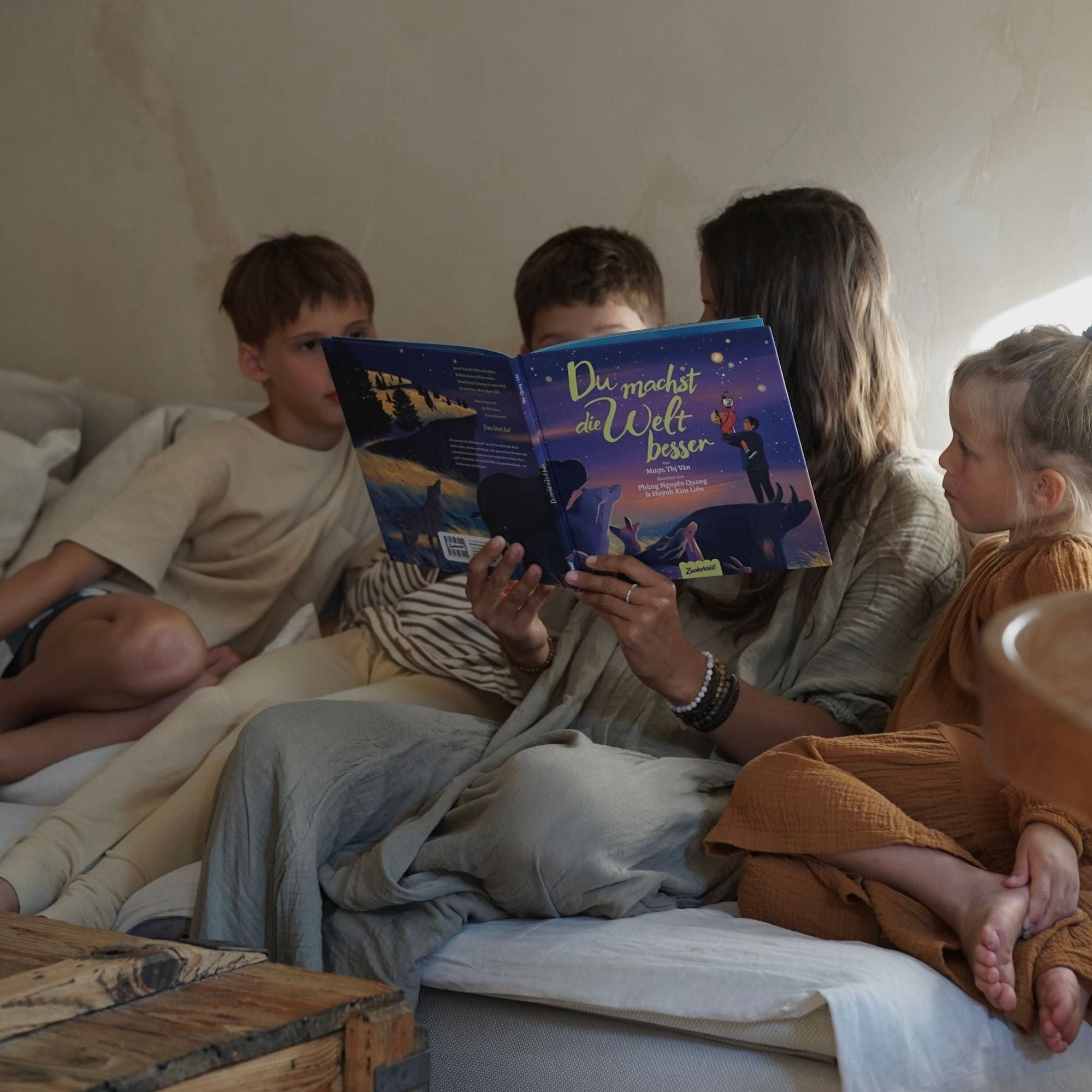 Eine Frau liest drei Kindern auf der Couch "Du machst die Welt besser" vom Zuckersüß Verlag vor und lädt sie in eine Welt der Zuversicht und des Staunens ein, während sie aufmerksam zuhören.