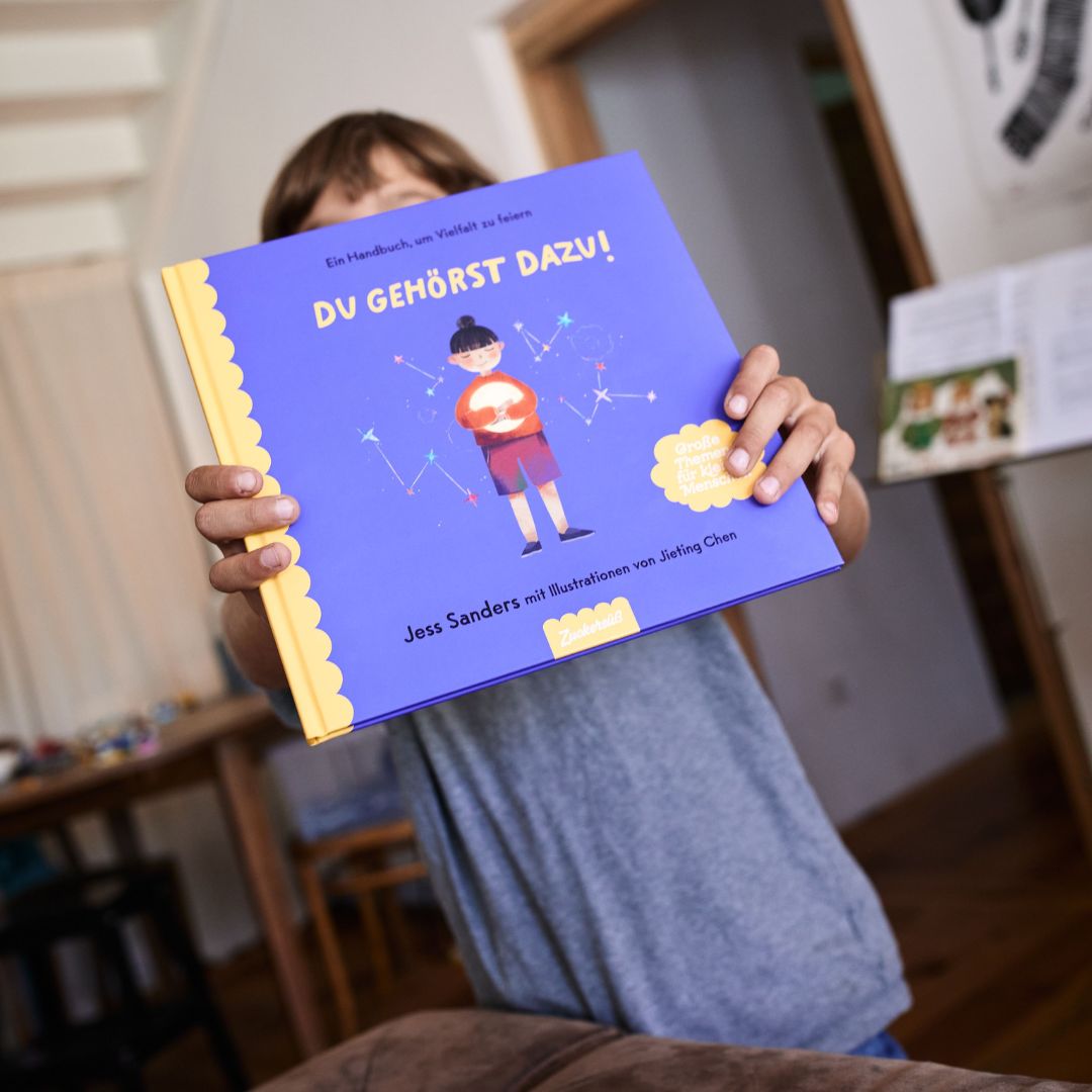 Ein Kind hält „Du gehörst dazu!“ hoch – ein deutsches Kinderbuch des Zuckersüß-Verlags, das sich der Vielfalt widmet – in einem gemütlichen Wohnzimmer, in dessen Hintergrund Möbel und eine Tür zu sehen sind.