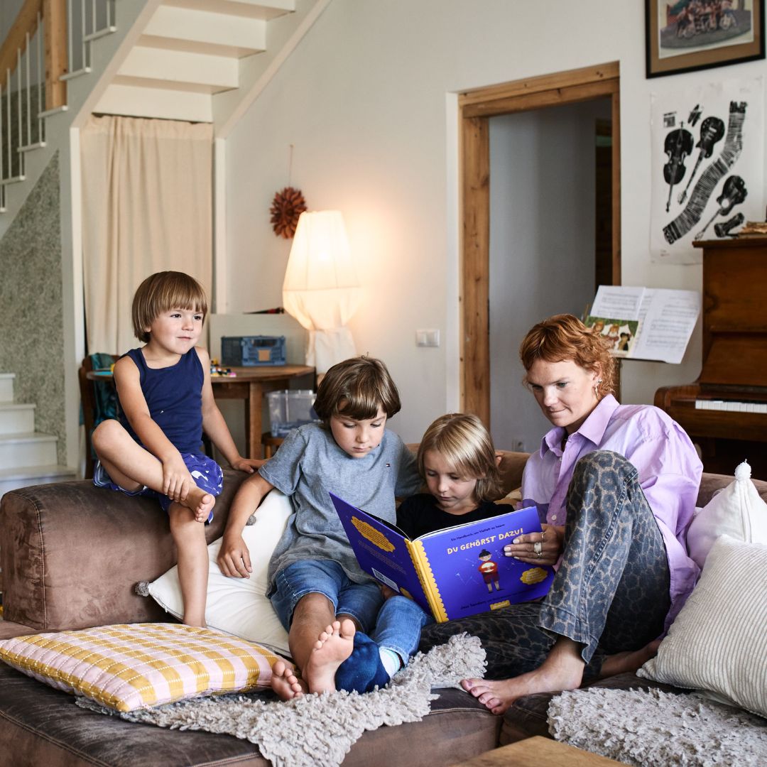 Eine Frau und drei kleine Kinder sitzen auf einem Sofa in einem gemütlichen Wohnzimmer und lesen gemeinsam das Buch „Du gehörst dazu!“ aus dem Zuckersüß-Verlag. Die Kinder sind ganz in die Geschichte vertieft, und im Zimmer stehen ein Klavier, eine Treppe und eine gemütliche Einrichtung.