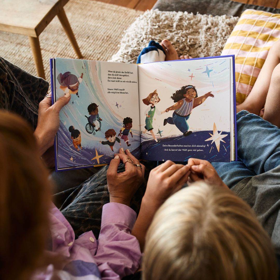 Zwei Menschen sitzen eng beieinander und lesen „Du gehörst dazu!“ – ein illustriertes Kinderbuch des Zuckersüß-Verlags, in dem auf bunten Seiten verschiedene Kinder zwischen Sternen herumlaufen und spielen. Das Bilderbuch liegt auf ihrem Schoß, während sie auf einem gemütlichen Sofa sitzen.