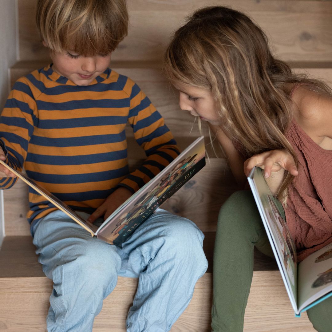 Zwei Kinder sitzen auf einer Holztreppe und lesen Diversität Kinderbücher. Das Mädchen, das grüne Leggings und ein ärmelloses Oberteil aus der Mädchen-Kollektion "Eine Welt wie diese" des Zuckersüß Verlags trägt, ist ganz auf ihr Buch konzentriert.