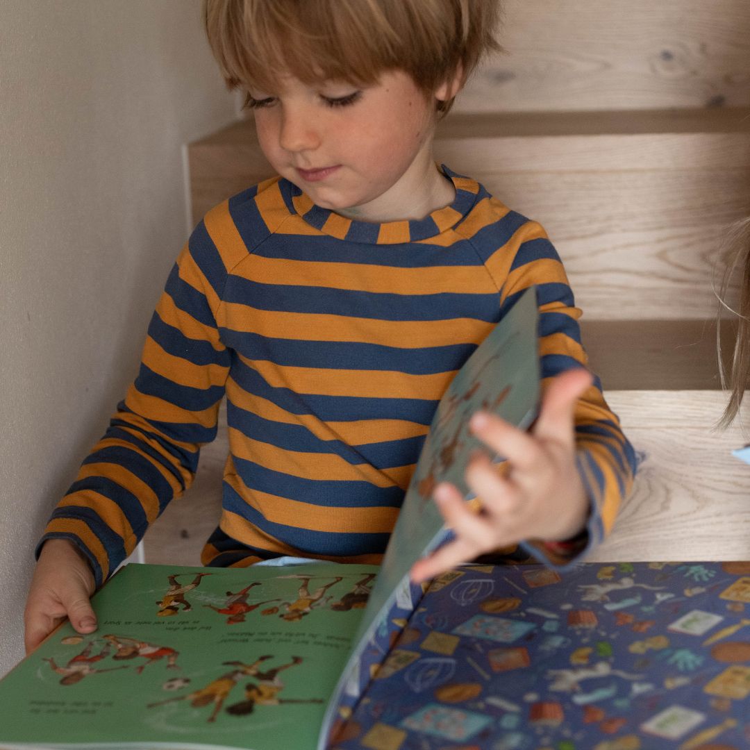Ein kleines Kind in einem blau-gelb gestreiften Hemd sitzt auf einer Holztreppe und schaut auf ein aufgeschlagenes Buch aus der "Eine Welt wie diese - Jungs-Kollektion" des Zuckersüß Verlags in seinem Schoß.
