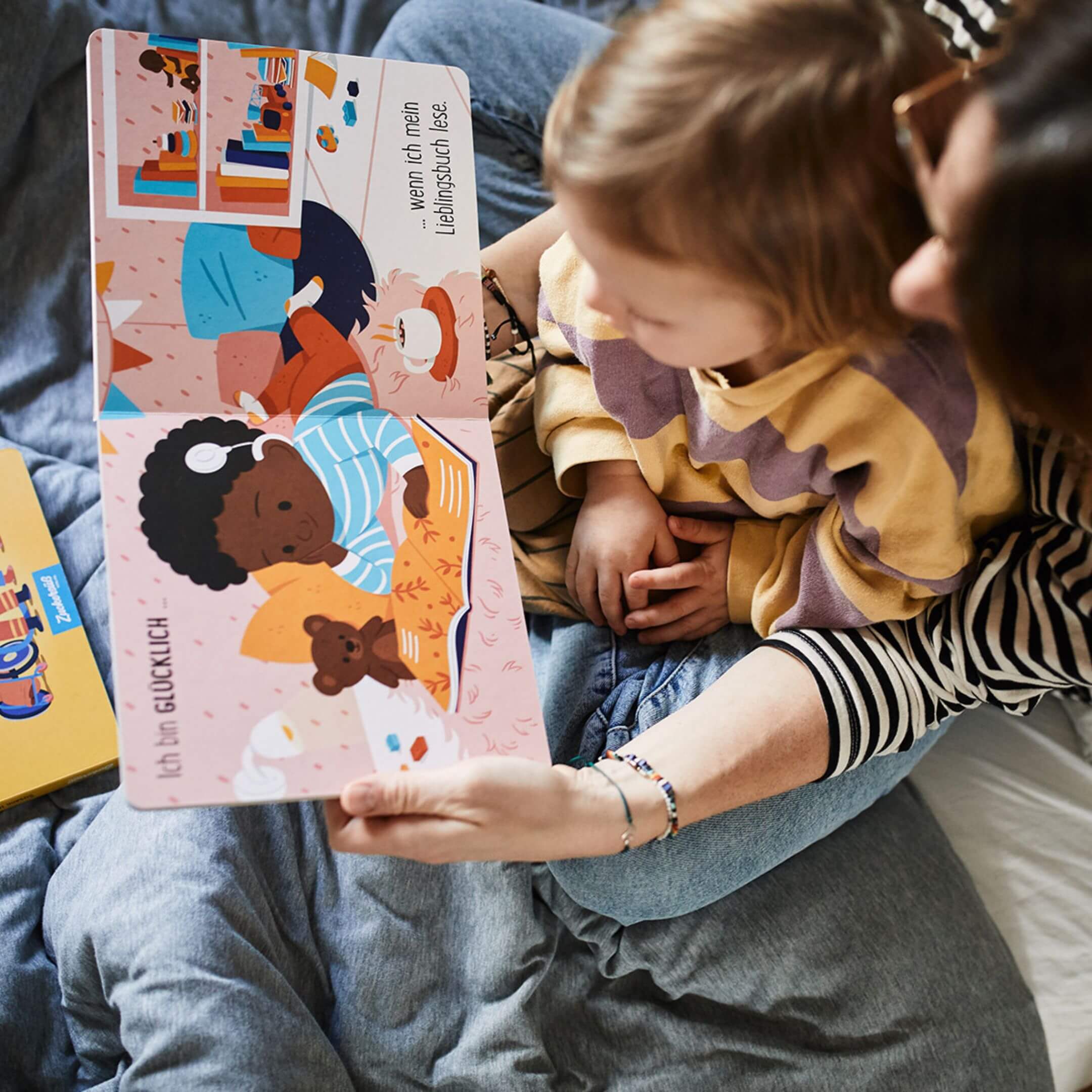 Ein Erwachsener und ein Kind sitzen eng beieinander auf einem Bett und schauen sich eine Doppelseite aus dem Pappbilderbuch „Gefühle sind für alle da!“ von Susann Hoffmann an. Die geöffnete Seite zeigt ein lesendes Kind mit dunkler Haut, Kopfhörern und Kuscheltier, daneben steht der Text: „Ich bin GLÜCKLICH … wenn ich mein Lieblingsbuch lese.“