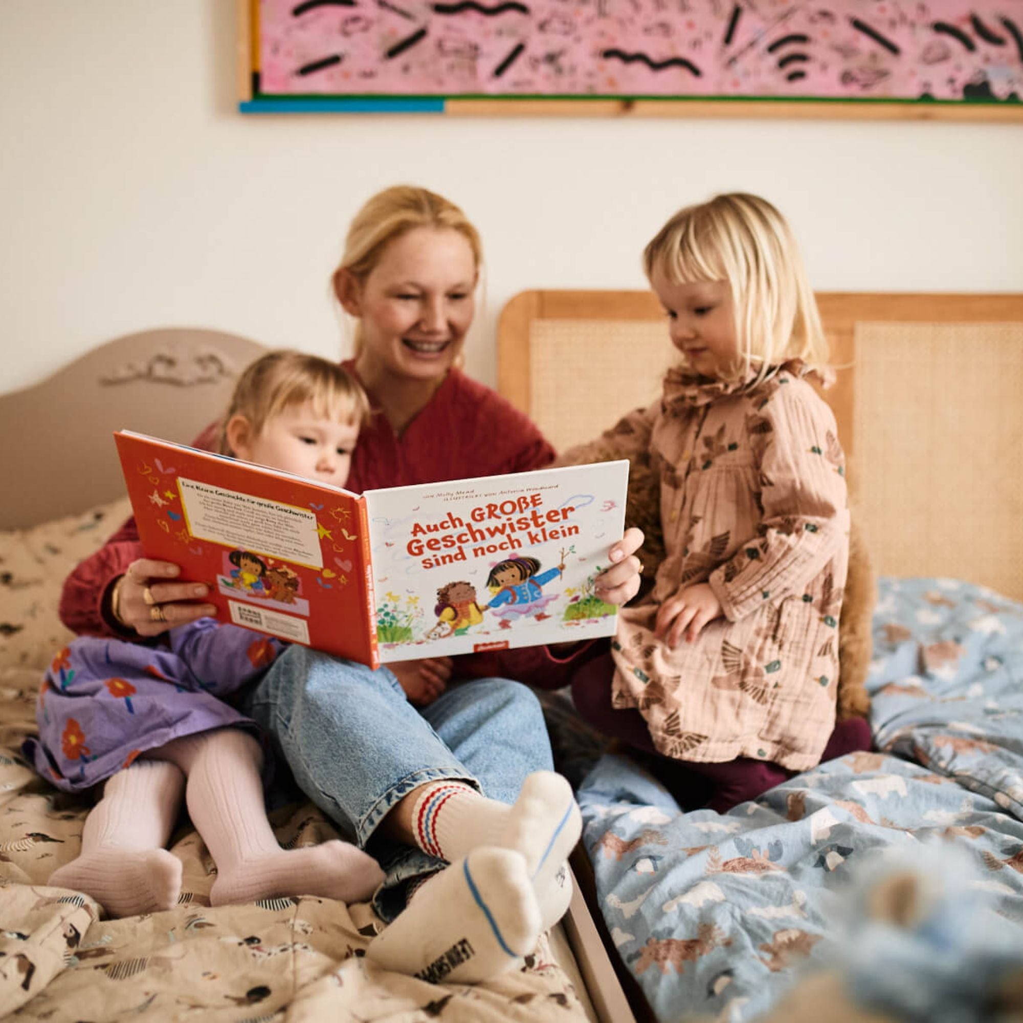 Eine Frau liest zwei Mädchen auf einem Bett "Auch große Geschwister sind noch klein" aus dem Zuckersüß Verlag vor. Sie lächeln, beschäftigt mit dem deutschen Bilderbuch über Gefühle, umgeben von farbenfrohem Dekor, das ihre geschwisterliche Verbundenheit unterstreicht.
