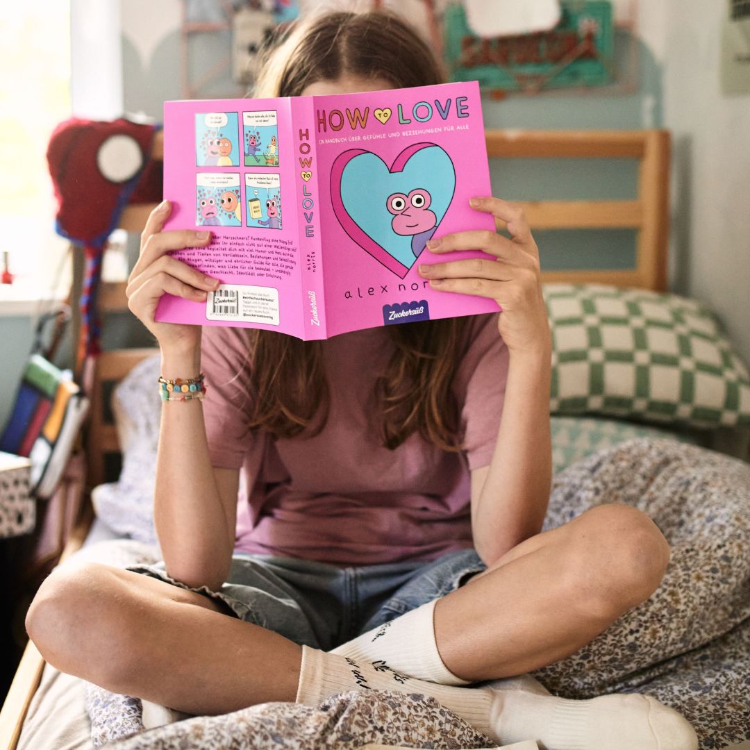 Eine Person sitzt mit gekreuzten Beinen auf einem Bett und hält das rosa gebundene Buch „How to love“ vom Zuckersüß Verlag hoch, wobei sie ihr Gesicht verbirgt. Der gemütliche Raum – akzentuiert durch gemusterte Kissen und Deko – schafft einen einladenden Rahmen, um sich mit Themen wie Liebe und Beziehungen auseinanderzusetzen.