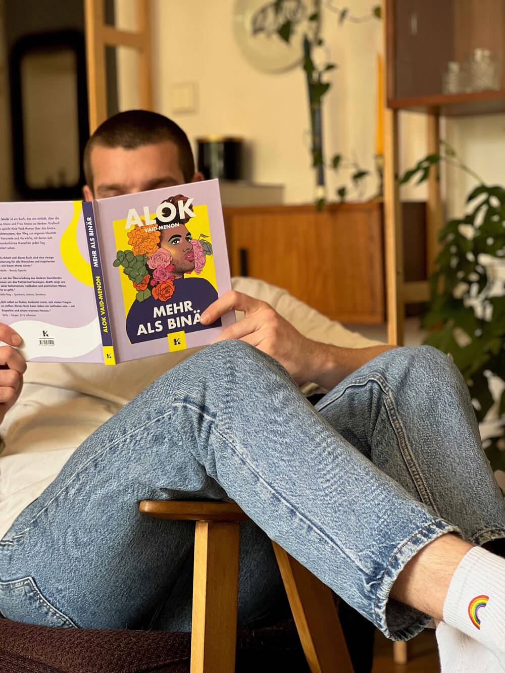 Eine Person entspannt sich mit gekreuzten Beinen und liest "Mehr als binär" von Alok Vaid-Menon. Sie ist mit hellblauen Jeans und regenbogenfarbenen Socken bekleidet und von Zimmerpflanzen und Holzmöbeln umgeben.