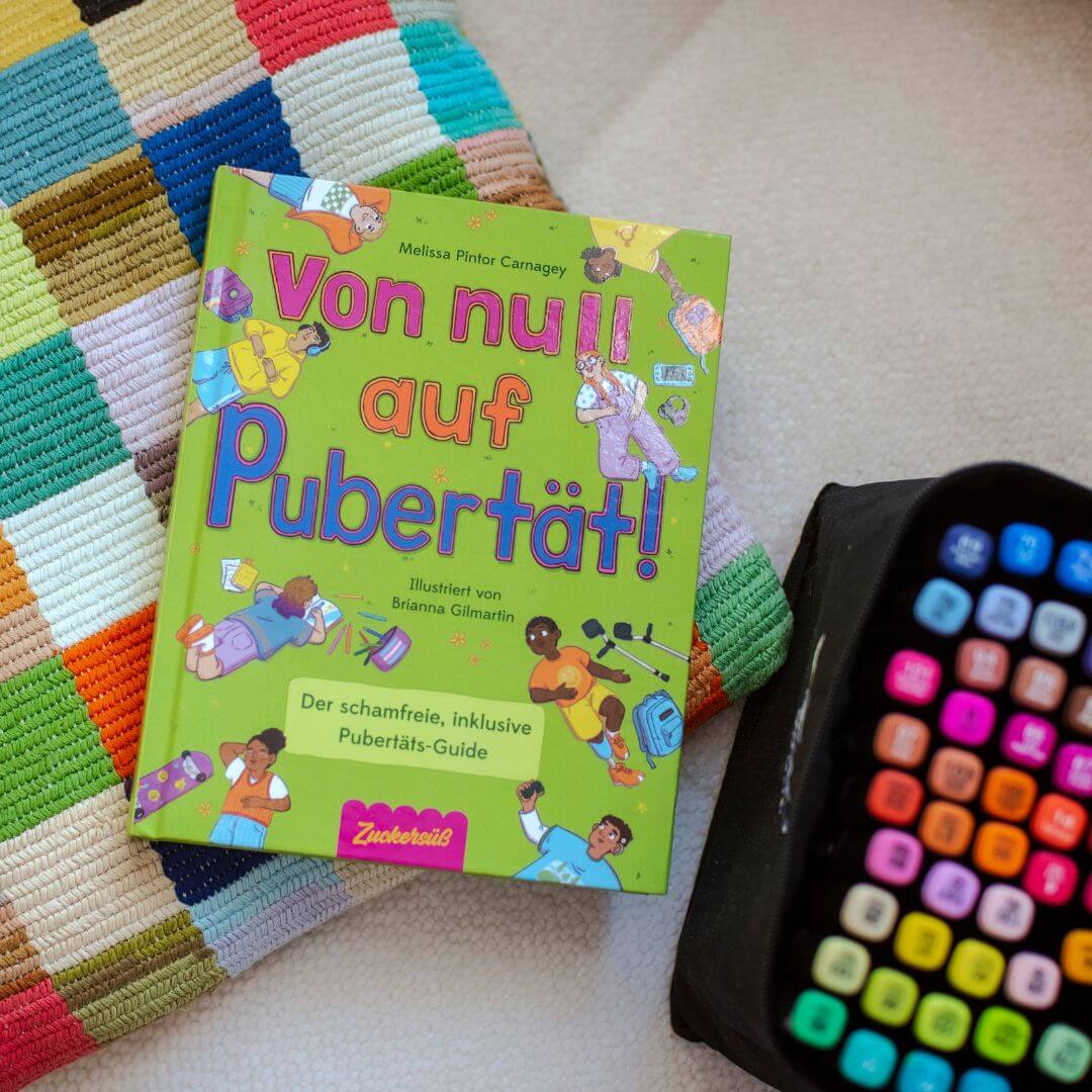 Das deutsche Buch "Von Null auf Pubertät" aus dem Zuckersüß Verlag liegt auf einer gestreiften Webmatte neben einer schwarzen TV-Fernbedienung. Sein verspieltes Cover behandelt Themen wie körperliche Veränderungen und psychische Gesundheit bei Kindern.