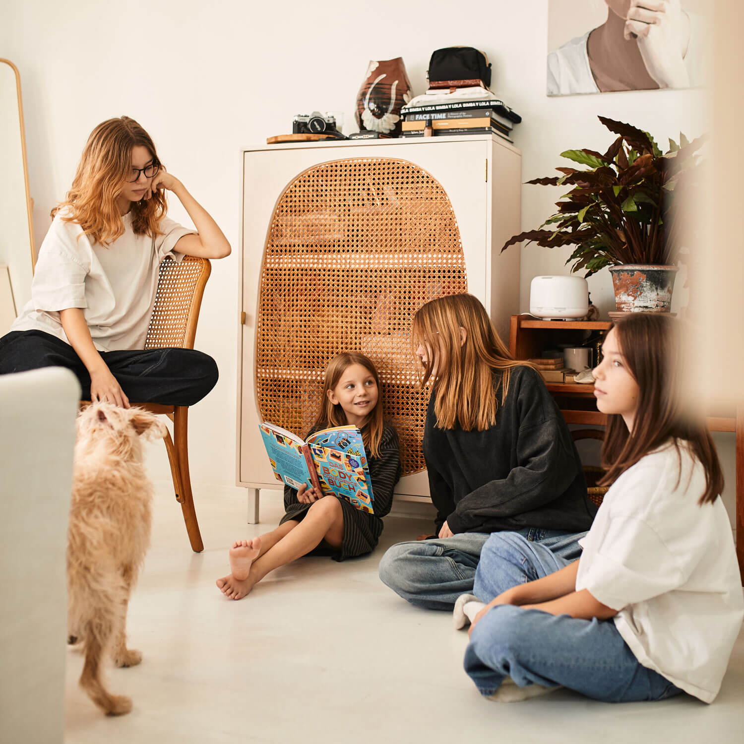Eine Frau sitzt auf einem Stuhl und streichelt einen Hund, während drei Kinder, von denen eines das "Das Body Confidence" Buch von Prof. Dr. Phillippa Diedrichs in der Hand hält, auf dem Boden in einem gemütlichen modernen Raum mit warmer Einrichtung und Pflanzen sitzen.