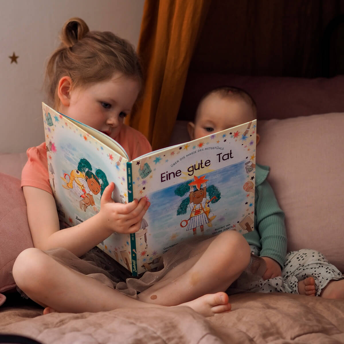 Ein junges Kind sitzt im Schneidersitz auf einem Bett und liest einem Baby neben sich "Eine gute Tat" von Christian Trimmer vor, beide umgeben von Kissen in einem warmen, gemütlichen Raum.