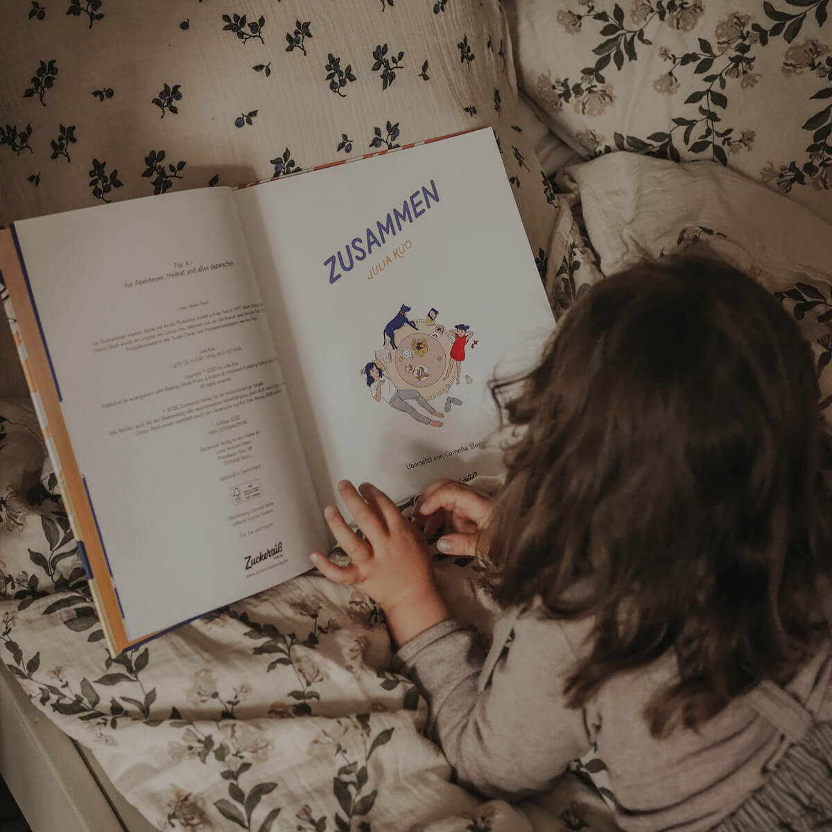 Ein Kind mit gewellten braunen Haaren liegt auf einem geblümten Bett und blättert im Bilderbuch „Zusammen“ von Julia Kuo. Das geöffnete Buch zeigt das Titelblatt mit einer Illustration von Menschen und Tieren, die um einen Tisch liegen – eine einladende Szene über Nähe und Gemeinschaft.