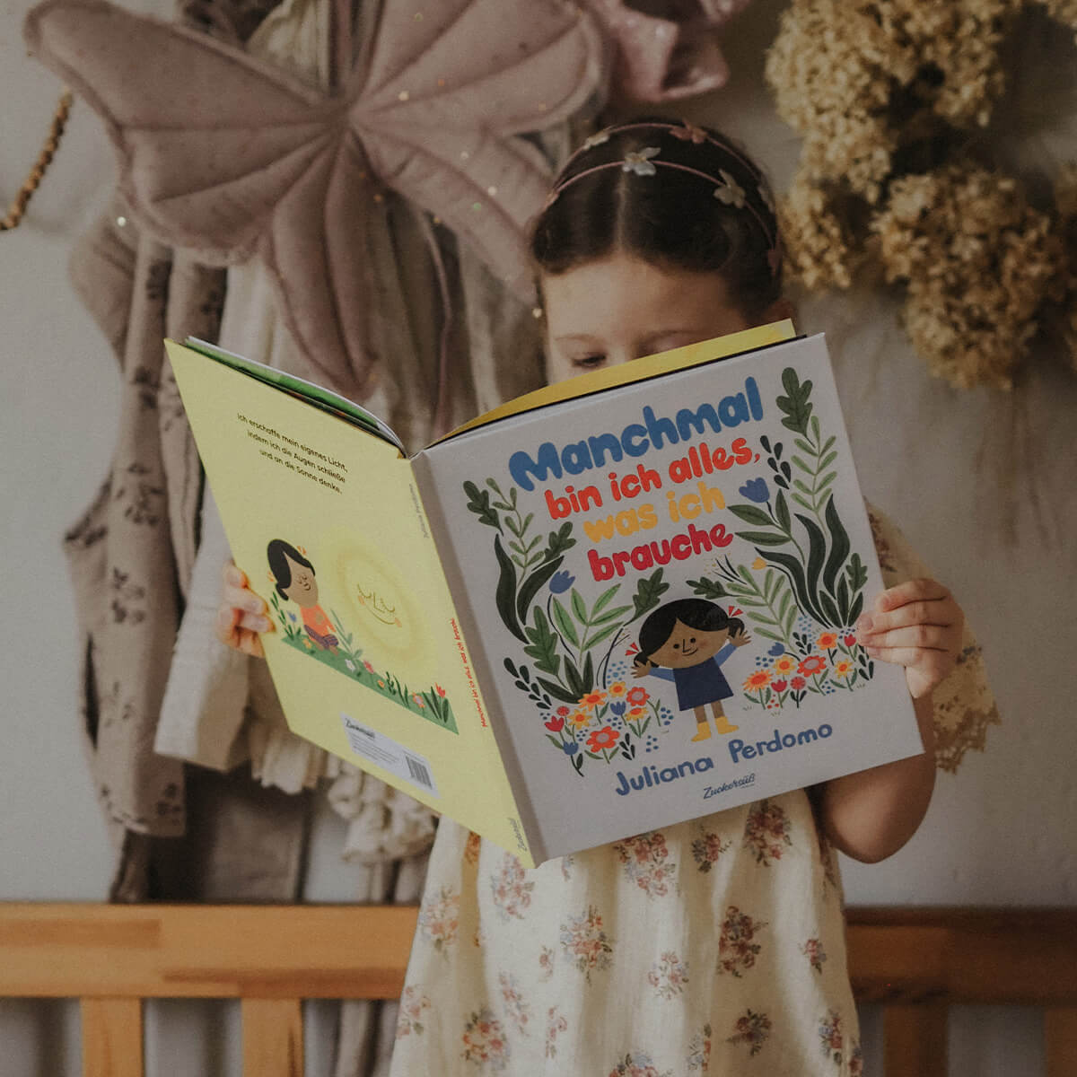 Ein Kind in einem geblümten Kleid steht vor einer Bank und hält das aufgeschlagene Bilderbuch „Manchmal bin ich alles, was ich brauche“ von Juliana Perdomo. Der farbenfrohe Buchumschlag mit Blumen und Pflanzenmotiven dominiert das Bild. Im Hintergrund hängen getrocknete Blumen und eine große Stoff-Schmetterlingsdekoration.