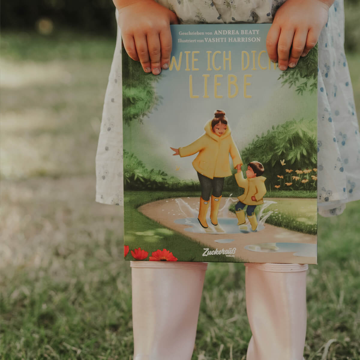Ein Kind in weißem Kleid mit blauen Punkten und rosa Strumpfhose steht auf einer Wiese und hält das Bilderbuch „Wie ich dich liebe“ von Andrea Beaty und Vashti Harrison in den Händen. Auf dem Cover springen ein erwachsener Mensch und ein Kind in gelben Regenmänteln fröhlich in eine Pfütze.