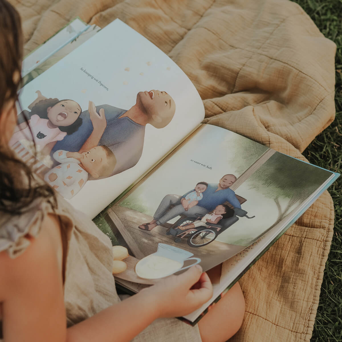Ein Kind sitzt auf einer beigefarbenen Decke im Gras und blättert im Bilderbuch Wie ich dich liebe von Andrea Beaty. Auf den offenen Seiten ist eine liebevolle Familienszene zu sehen: ein Vater im Rollstuhl sitzt mit zwei Kindern unter einem Baum, daneben eine Szene mit drei lachenden Kindern und dem Vater.