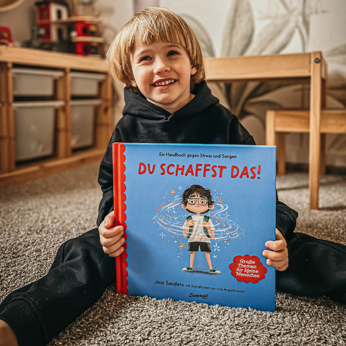 Ein lächelndes Kind in einem schwarzen Kapuzenpulli sitzt auf einem Teppichboden und hält das Buch "Du schaffst das!" von Jessica Sanders in der Hand, dessen Cover mit einem Kind illustriert ist. Im Hintergrund sind Holzmöbel zu sehen.