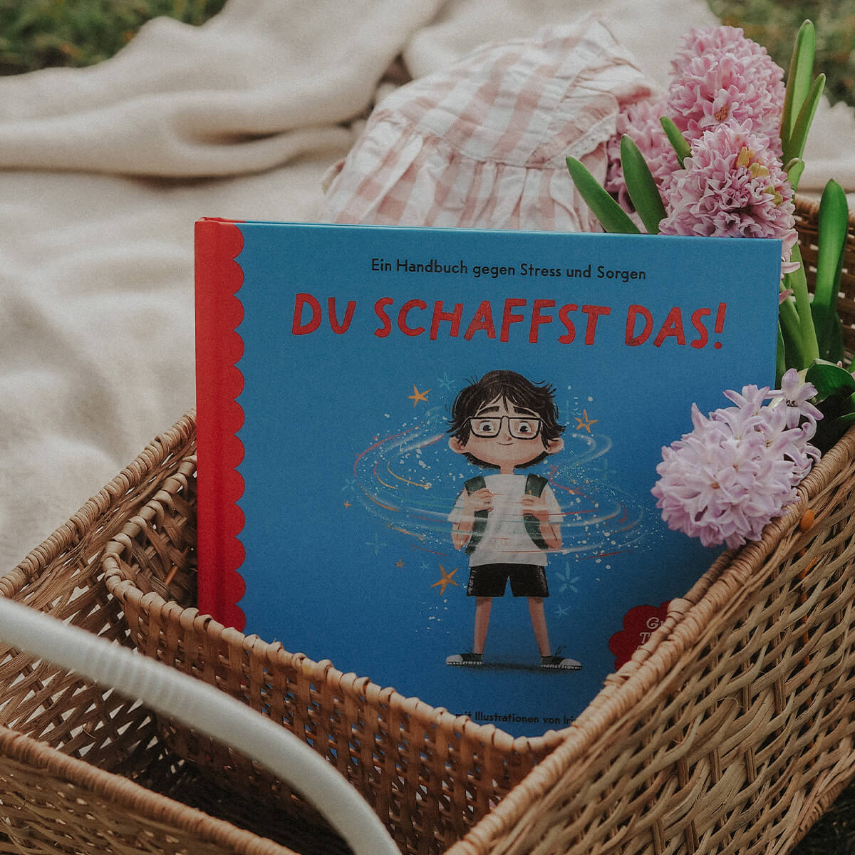 Das Kinderbuch "Du schaffst das!" von Jessica Sanders liegt in einem geflochtenen Korb neben rosa Hyazinthen auf einer Picknickdecke.