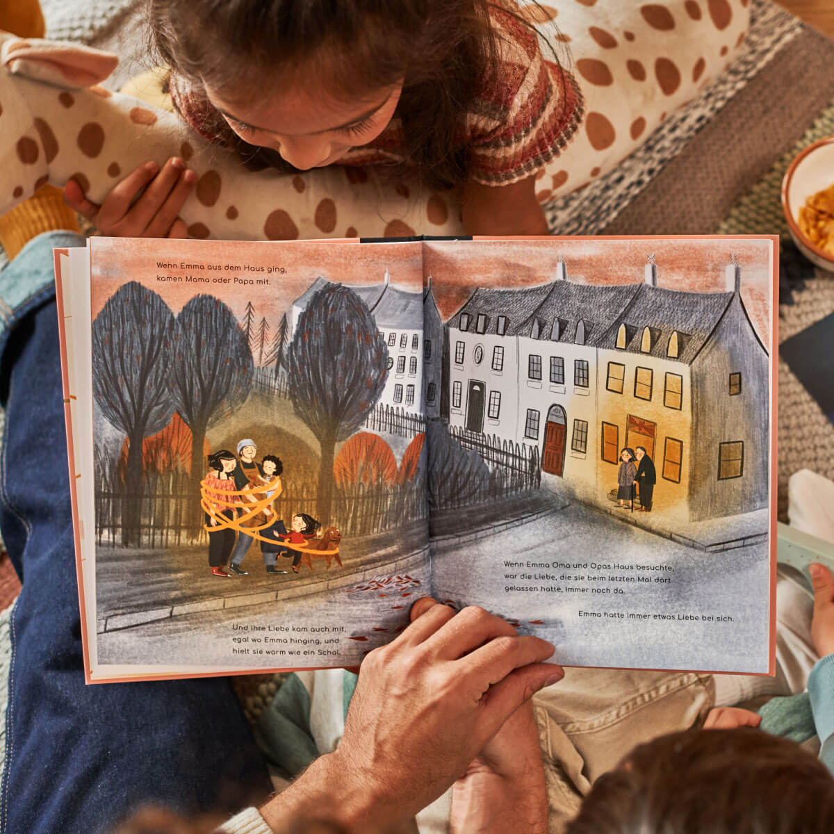 Ein Kind und ein Erwachsener sitzen zusammen und lesen "Liebe" von Corrinne Averiss. Die aufgeschlagenen Seiten zeigen farbenfrohe Illustrationen von Familien, die eine von Bäumen gesäumte Straße entlang spazieren, während ihre Hände beim Lesen auf dem Buch ruhen.