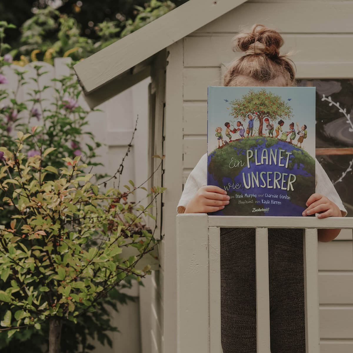 Auf der Veranda eines kleinen hölzernen Spielhauses hält sich ein Kind "Ein Planet wie unserer" von Frank Murphy & Charnaie Gordon vor das Gesicht, wobei eine Blattpflanze in der Nähe auf das Interesse am Umweltschutz hindeutet.