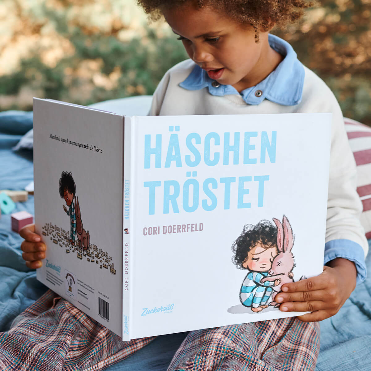 Ein Kind sitzt draußen auf einer Decke und liest Häschen tröstet aus dem „Best of Cori Doerrfeld“-Paket vom Zuckersüß Verlag. Das illustrierte Cover zeigt ein Kind, das ein Häschen umarmt.