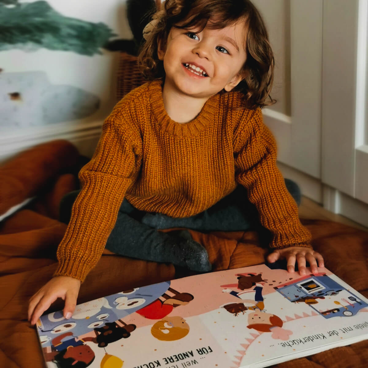 Ein lächelndes Kind mit gewellten braunen Haaren und einem senfgelben Pullover sitzt auf einer braunen Decke und blickt auf, während es das bunte Pappbilderbuch "Spielzeug ist für alle da!" von Susann Hoffmann in der Hand hält.