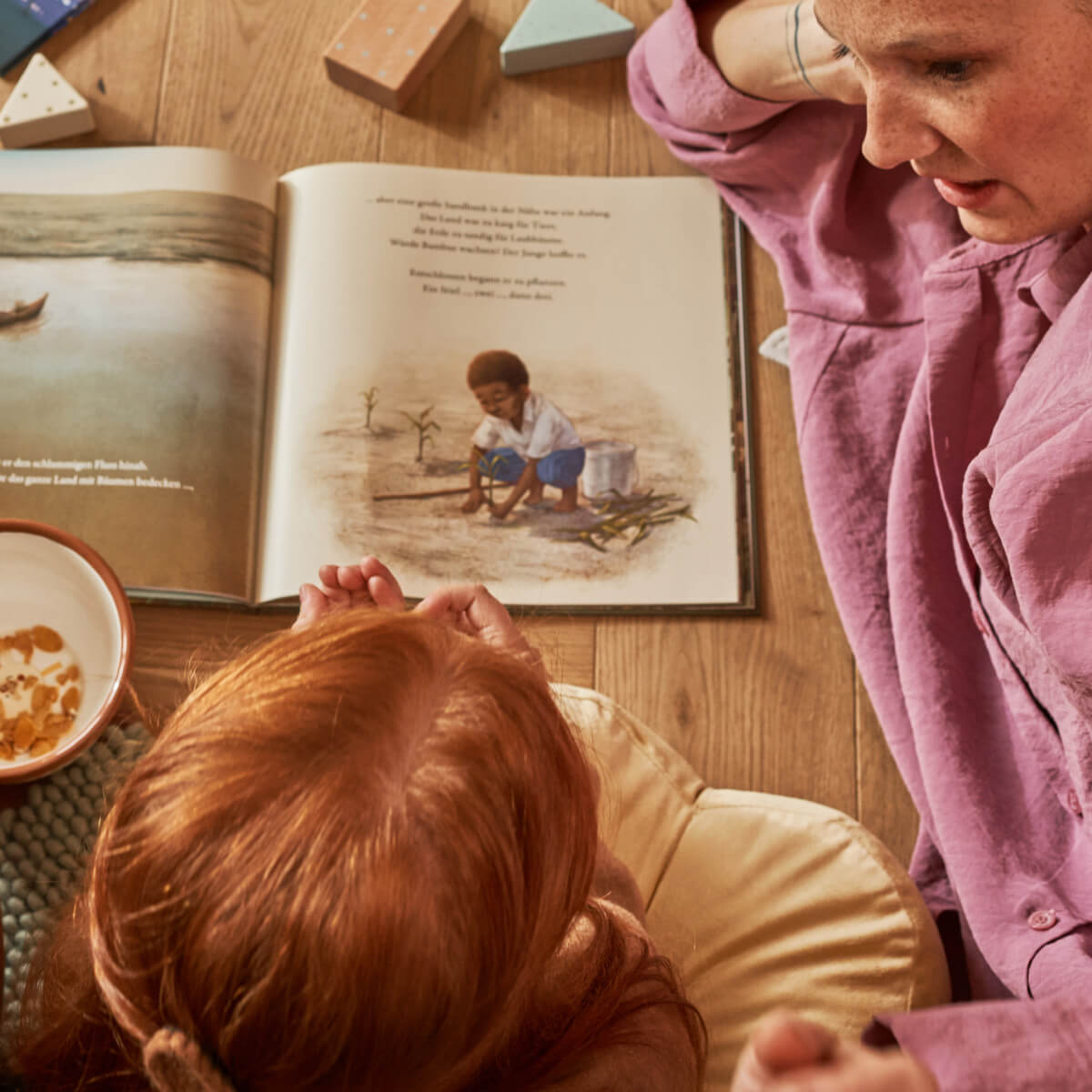 Ein Erwachsener und ein rothaariges Kind sitzen auf einem Holzboden und lesen "Der Junge, der einen Wald pflanzte" von Sophia Gholz. Die aufgeschlagene Seite zeigt ein Kind bei der Arbeit am Wasser. In der Nähe steht eine Schale mit Müsli.