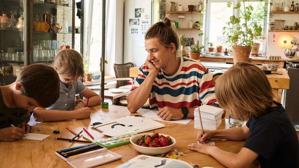 Eine erwachsene Person sitzt mit drei Kindern am Tisch und lächelt. Die Kinder malen