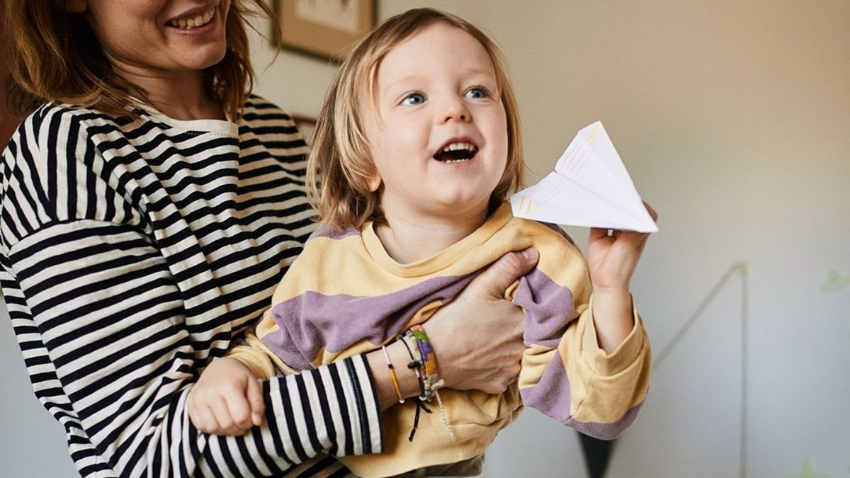 ein Kind im Arm seiner Mutter hält einen Papierflieger in der Hand und lacht