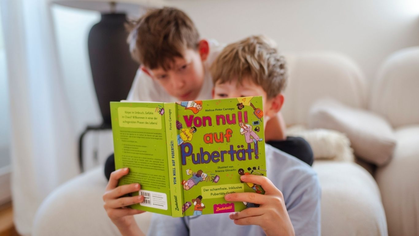 zwei Jungs sitzen auf der Couch und lesen gemeinsam "Von null auf Pubertät" vom Zuckersüß Verlag