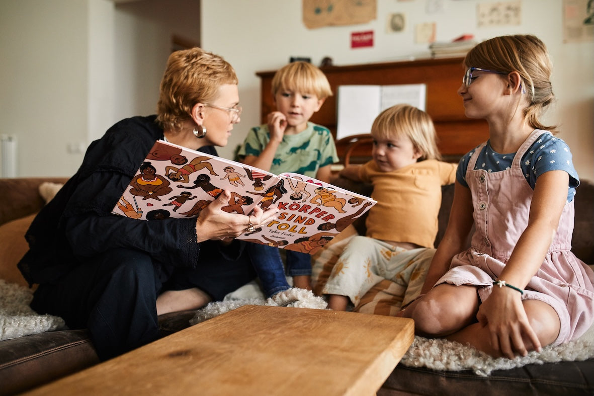 Ein Erwachsener liest drei aufmerksamen Kindern auf einer gemütlichen Wohnzimmercouch "Das Kennlern-Paket" aus dem Zuckersüß Verlag vor - eines der meistverkauften Kinderbücher über Body Positivity.