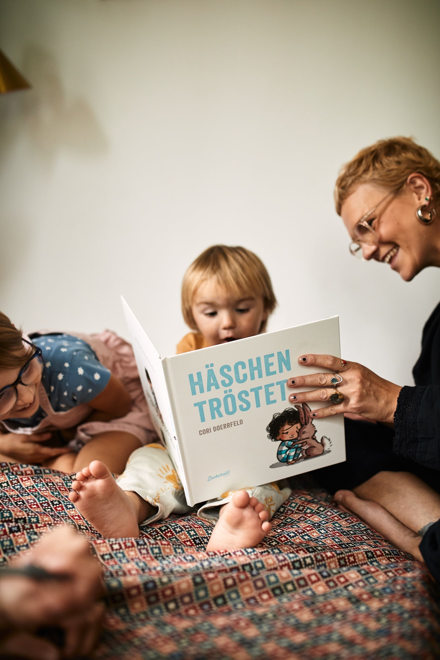 Ein Erwachsener und zwei Kinder sitzen auf einer gemusterten Bettdecke und machen es sich beim Lesen von Das Kennlern-Paket aus dem Zuckersüß Verlag, einem der meistverkauften Bücher in Deutschland, gemütlich. Die Kinder sehen beschäftigt und glücklich aus.