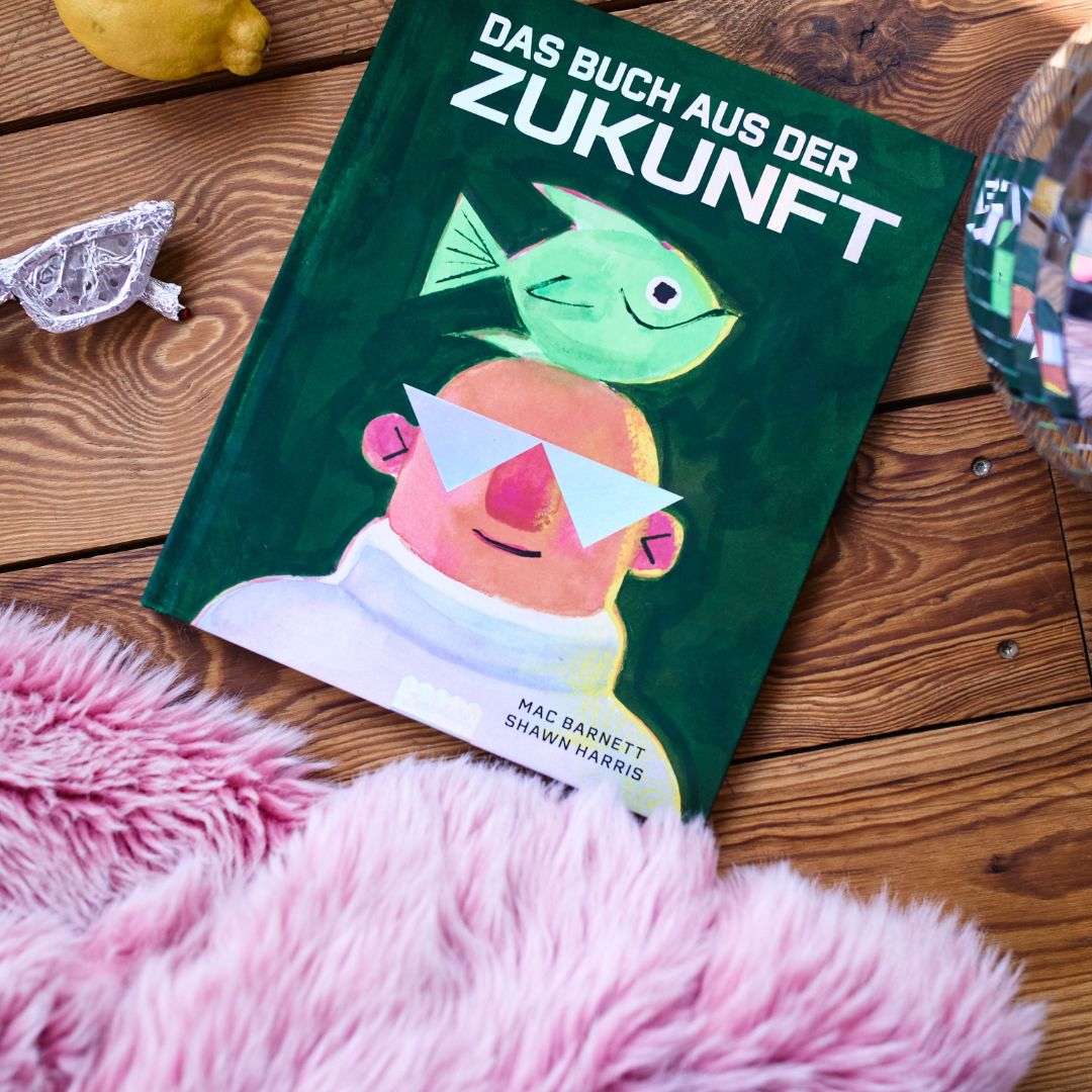 Das Buch des Zuckersüß-Verlags mit dem Titel „Das Buch aus der Zukunft“, auf dessen Cover eine Comic-Illustration einer Person mit Sonnenbrille und einem Fisch auf dem Kopf zu sehen ist, liegt auf einem Holzboden neben einem rosa Plüschteppich, einer Zitrone, einer Glasfigur und einer Discokugel.