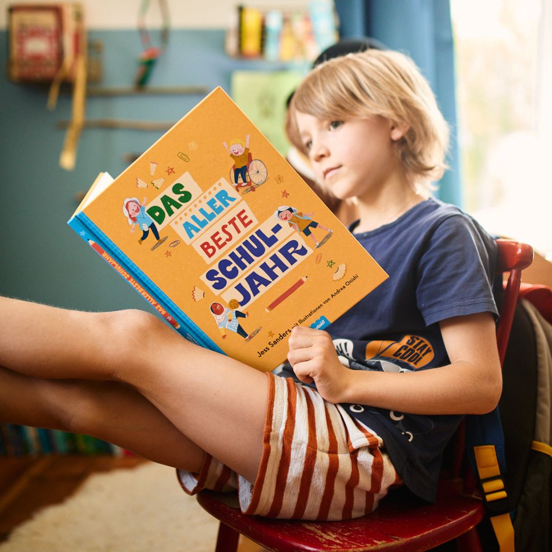 Ein blonder Junge, der entspannt auf einem roten Stuhl sitzt und ein Bein hochgelegt hat, liest in gestreiften Shorts und einem dunkelblauen T-Shirt das Buch „Das allerbeste Schuljahr“ vom Zuckersüß Verlag und genießt seine Schulabenteuer.