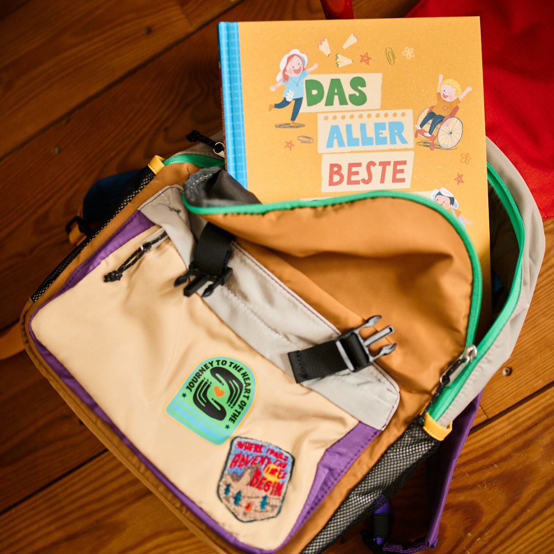 Ein Exemplar von „Das allerbeste Schuljahr“ vom Zuckersüß Verlag lugt aus einem bunten, mit Aufnähern verzierten Rucksack hervor – ideal für den Schulalltag –, der auf einem Holzboden steht.