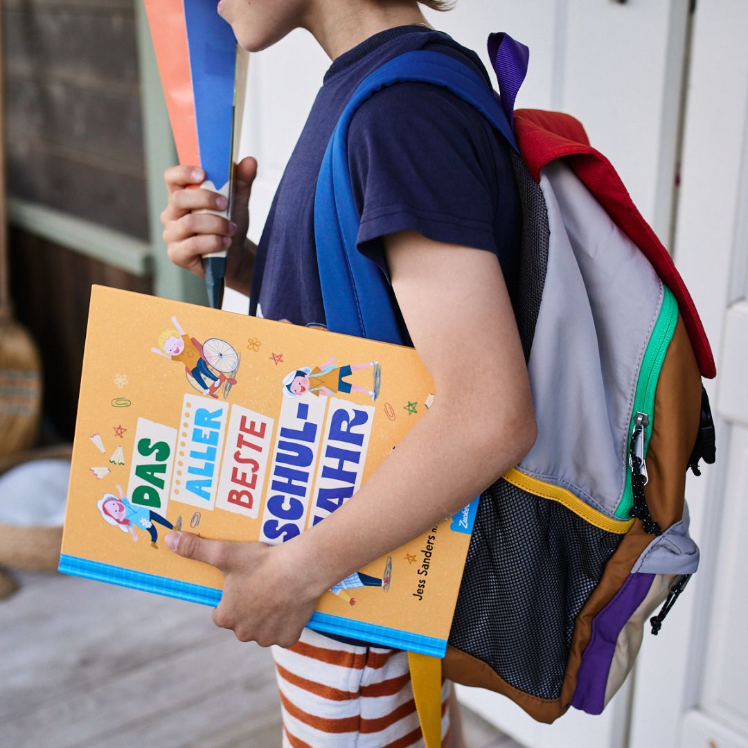 Ein Kind mit Rucksack steht vor einer gestreiften Tür und hält das Buch „Das allerbeste Schuljahr“ vom Zuckersüß Verlag in der Hand – voller bunter Illustrationen und perfekt für ein weiteres spannendes Schulabenteuer.